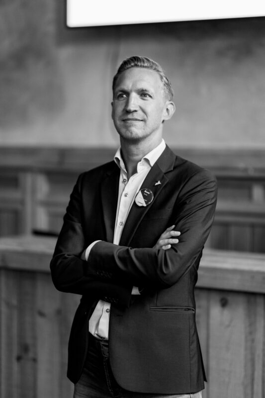 Portrait of Bruno Braes in a suit with arms crossed, smiling confidently in a professional indoor setting.