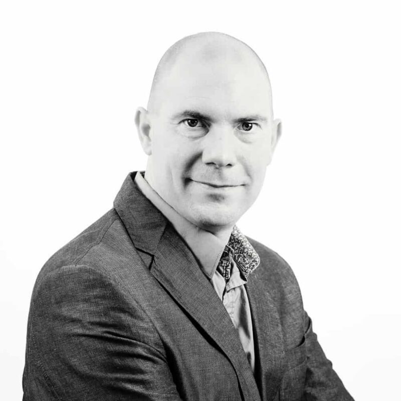 Portrait of Jo Putzeys smiling, wearing a suit jacket and collared shirt, facing forward on a white background.