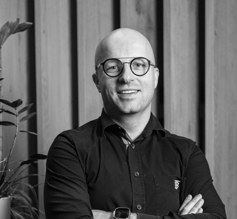 Portrait of Kevin van der Straeten smiling, wearing glasses and a dark shirt, standing in front of wooden panels.
