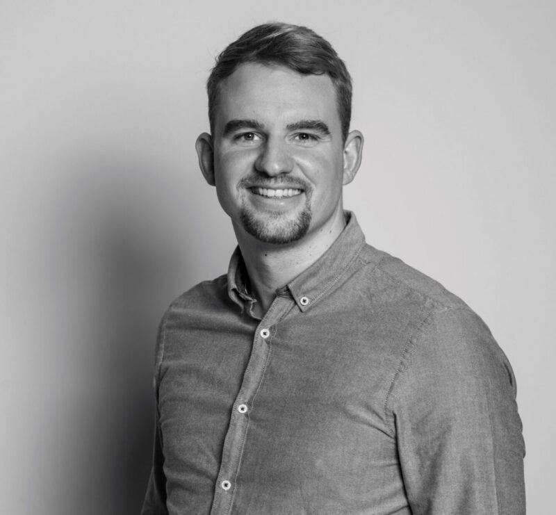 Portrait of Nick Windey smiling, wearing a button-up shirt, standing in front of a plain background.