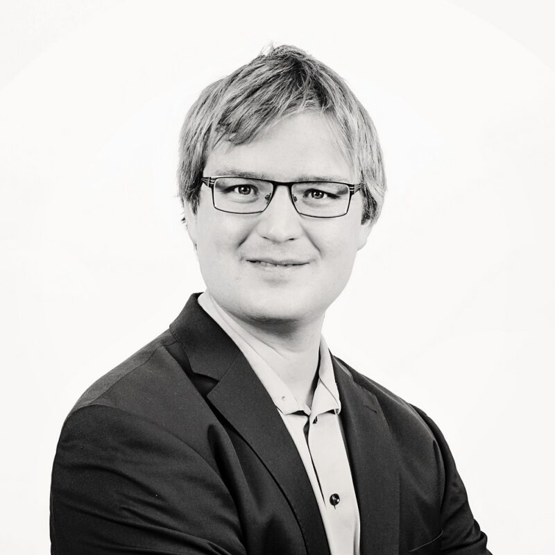 Portrait of Nicolas Delahaut smiling, wearing glasses and a suit jacket, in front of a white background.