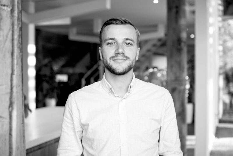 Portrait of Nathan Mollema smiling, wearing a light shirt, standing in a modern office interior.