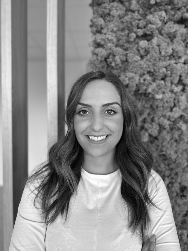 Portrait of Priscilla Calemi smiling, with long wavy hair, wearing a light top, in a modern indoor setting.