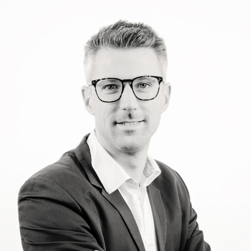 Portrait of Sam Geeraerts smiling, wearing glasses, a blazer and white shirt, on a white background.