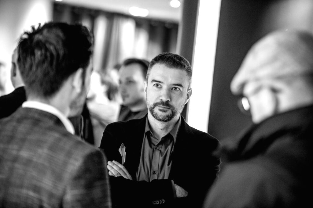 Tom Stroobants at a networking event, standing with arms crossed, in conversation with other attendees.