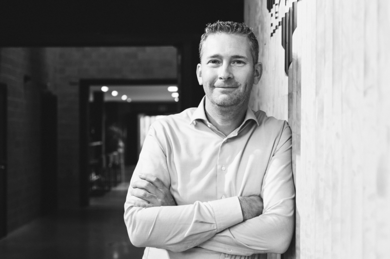 Tommy De Haan leans casually against a wall, smiling, arms crossed, in a modern hallway with ceiling lights.